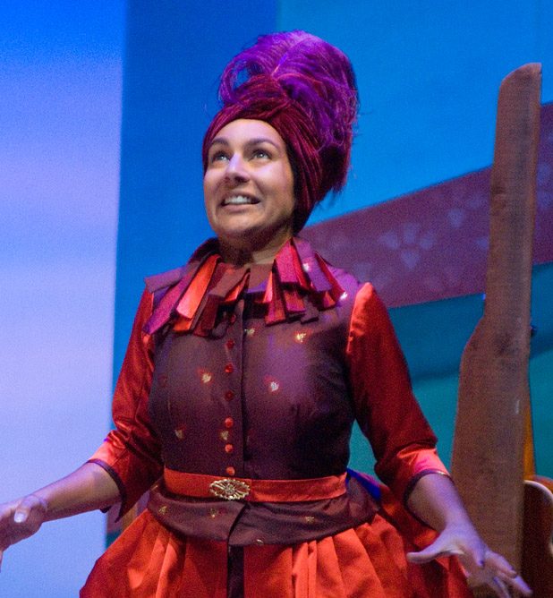 A brown-skinned woman in a two-toned red dress with a fitted bodice. She wears a feathered headress and is looking up excitedly.